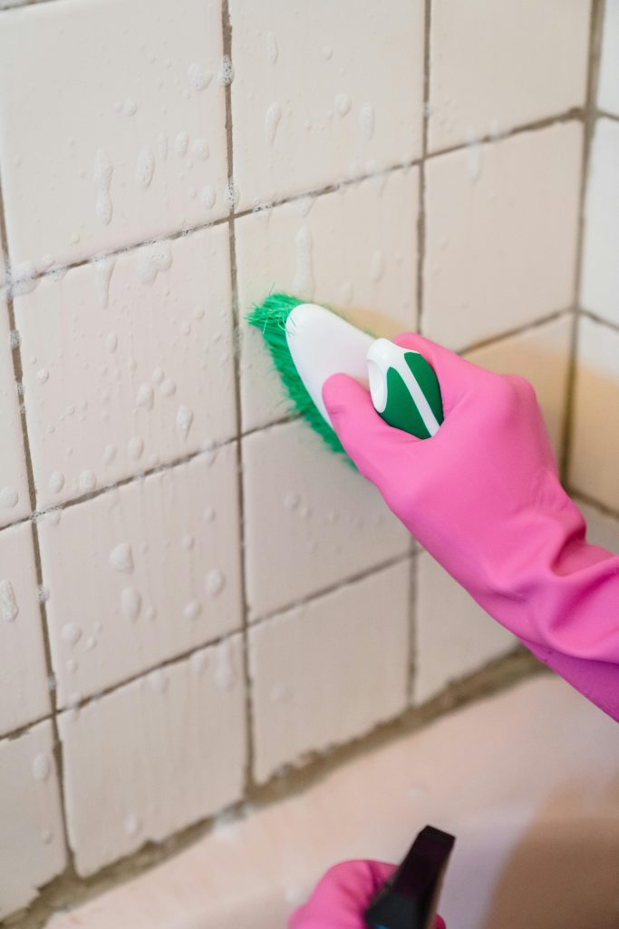 pexels-photo-9462638-9462638 A hand in pink gloves scrubs bathroom tiles with a green brush to remove grime.